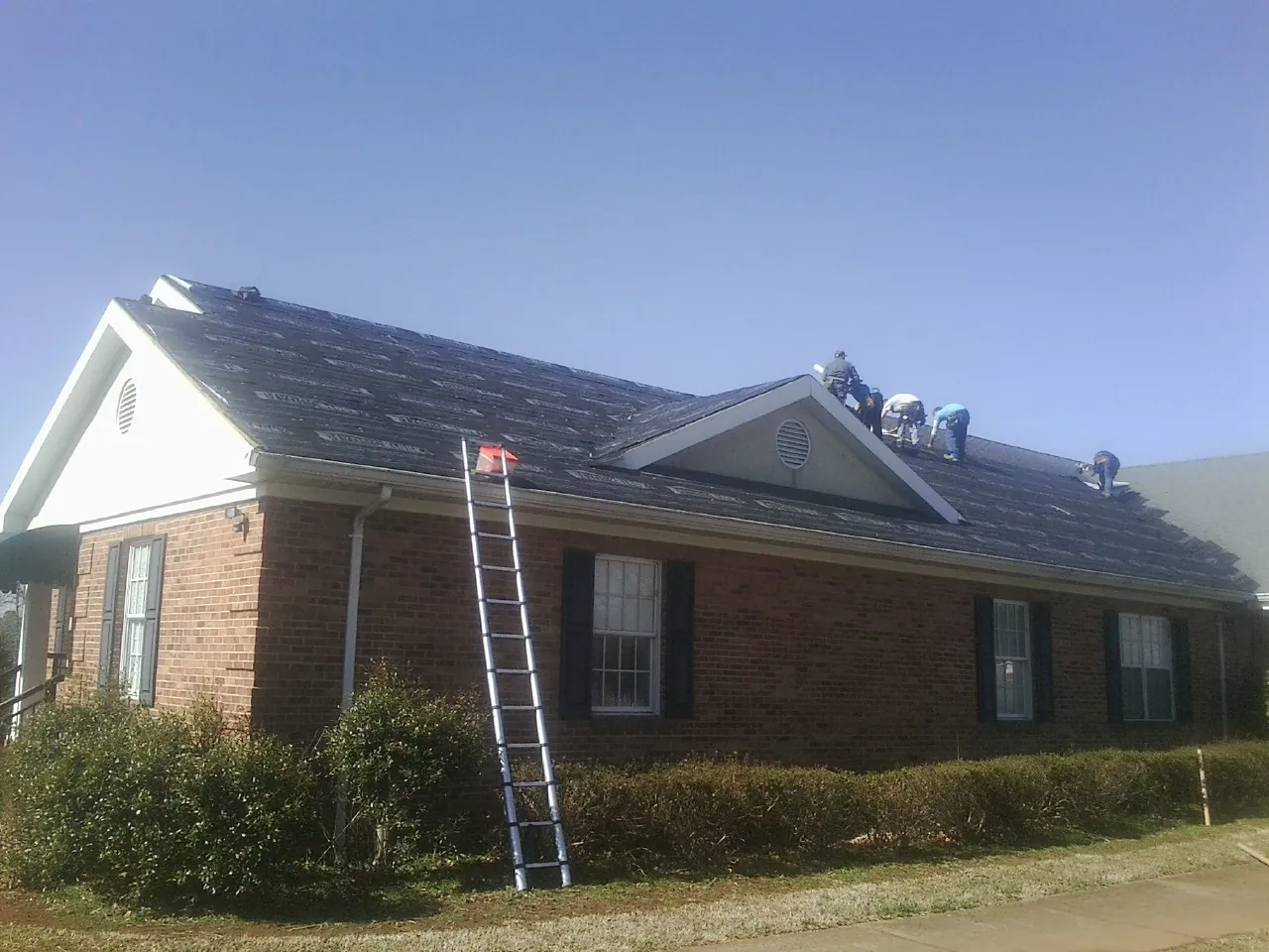Crew tearing shingles off a brick ranch roof with a ladder set — Emergency Roof Repair work by our crew in Middle Grove