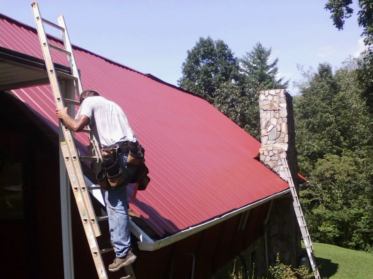 Roofer on a ladder beside a red metal roof and stone chimney — Soffit & Fascia Repair by Roofing Brothers in Middle Grove
